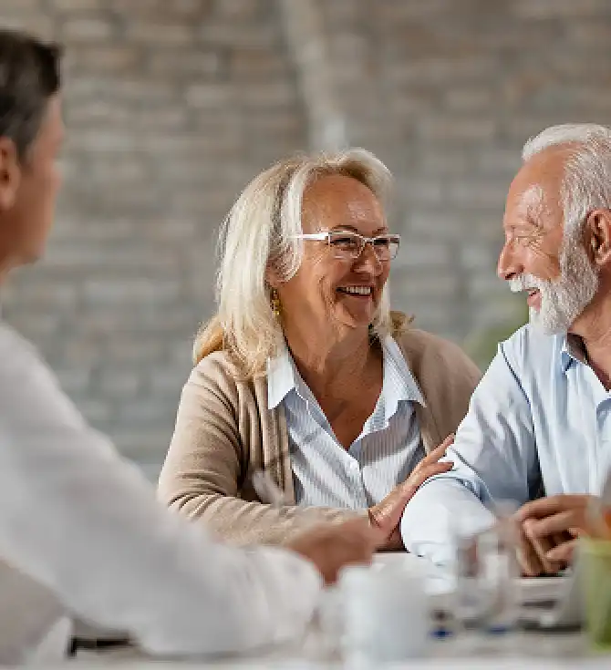 Three adults sit at a table indoors, smiling and chatting; a blond person with glasses and a white-haired person with a beard are in focus.