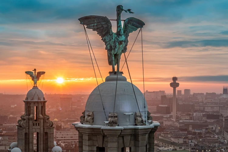 Two winged statues atop domed towers overlook a city at sunset, with orange horizon and blue sky over a dense urban landscape.